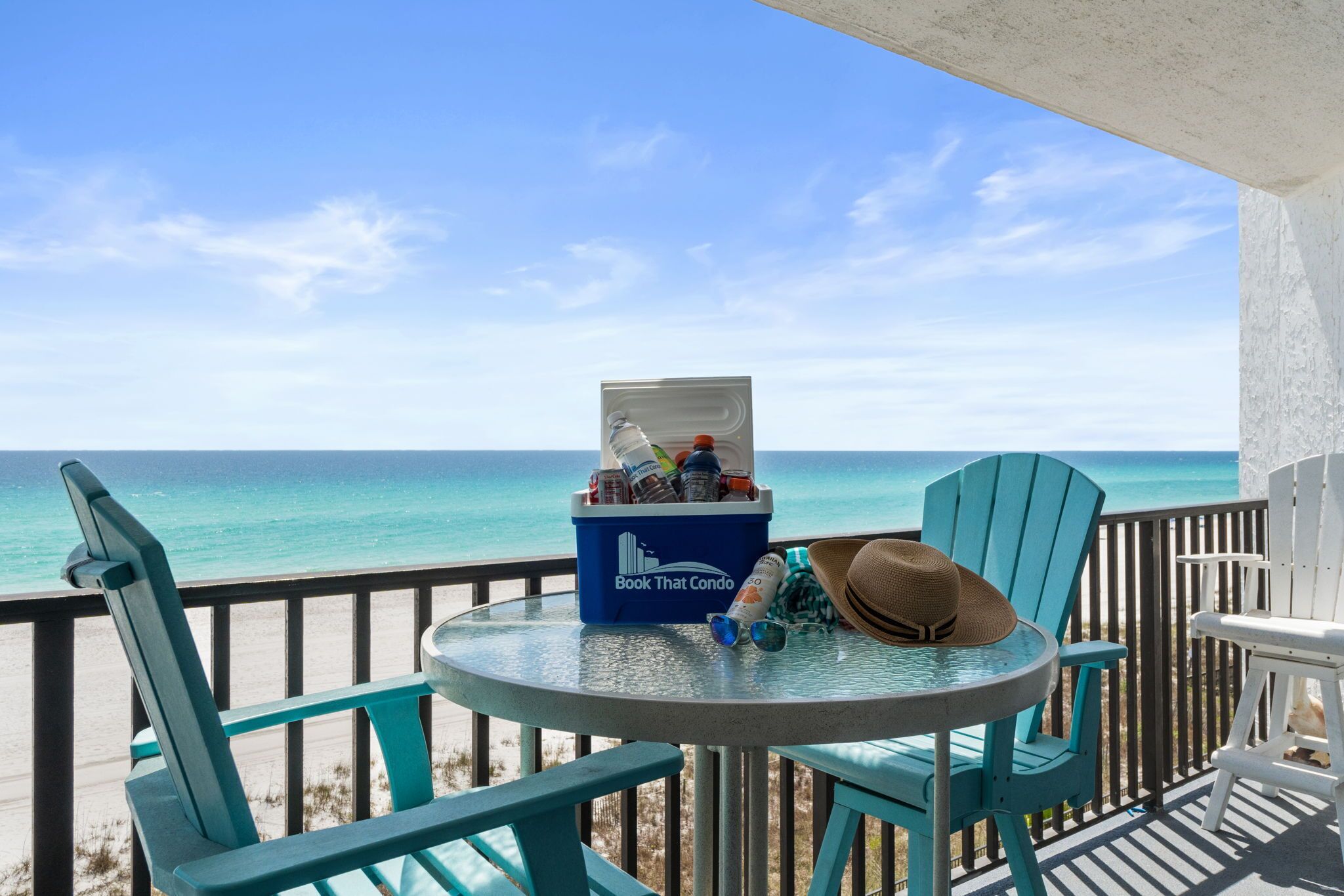 A balcony with a view of the ocean, a blue cooler labeled 'Book That Condo,' and turquoise chairs.