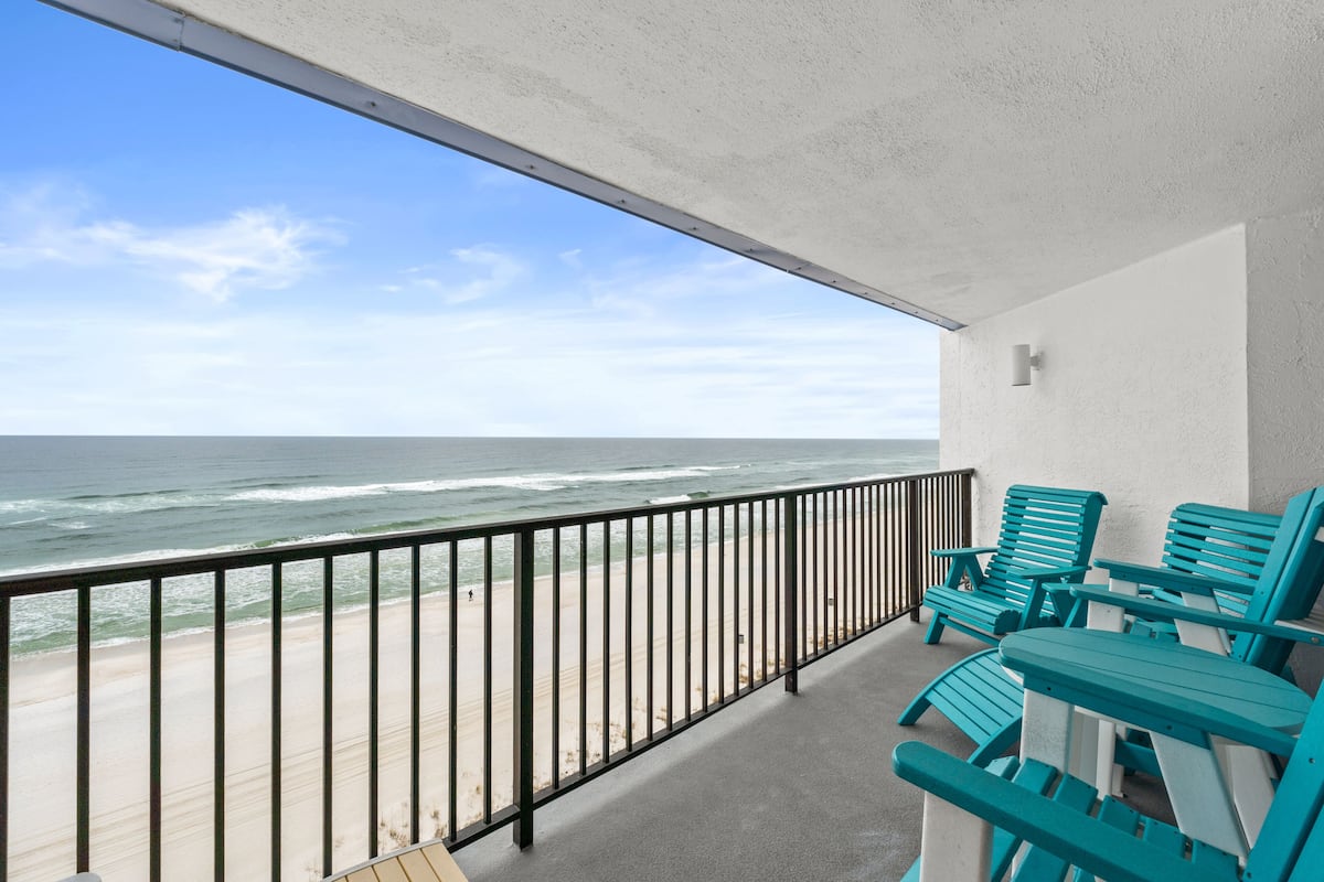 A balcony with a view of the ocean, featuring a railing and two turquoise Adirondack chairs.