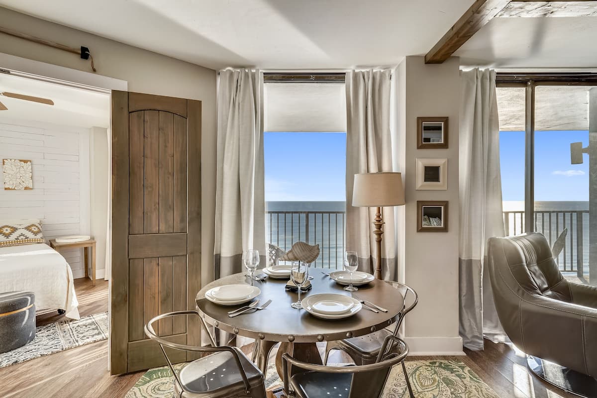 A dining area with a round table set for four, a view of the sea through large windows, and a balcony with a view.