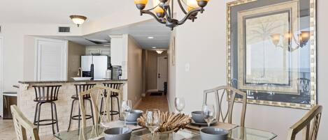 A dining area with a glass table, chairs, and a chandelier. There is a kitchen in the background.
