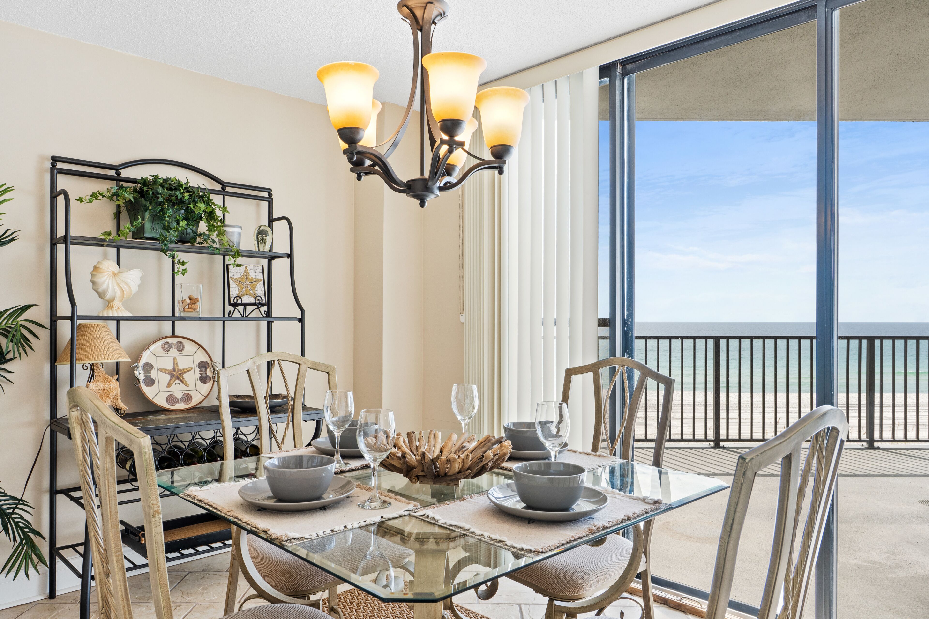 A dining area with a glass table, chairs, and a chandelier. There is a plant on a metal shelf and a view of the ocean through large windows.