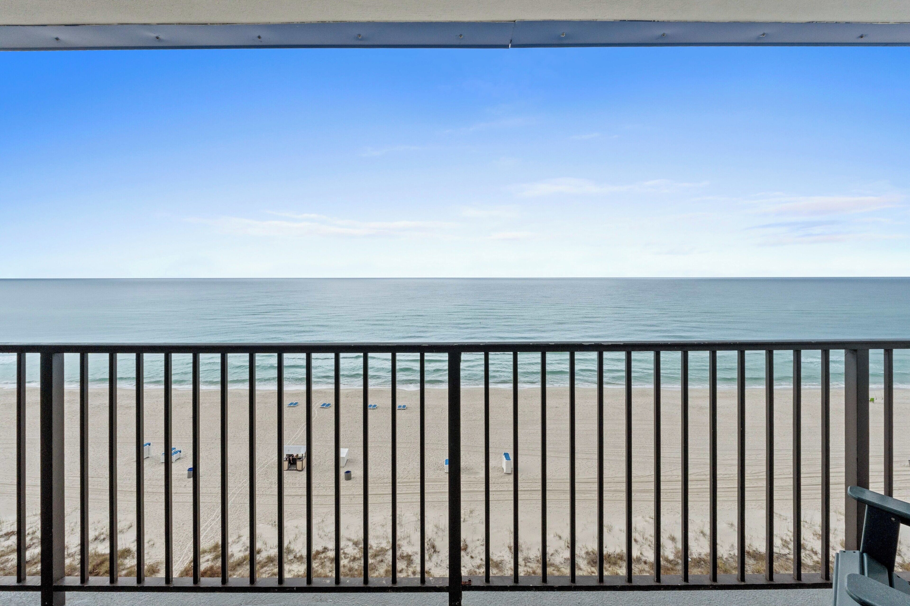 Vista a la playa desde un balcón con barandilla, cielo despejado y mar tranquilo.