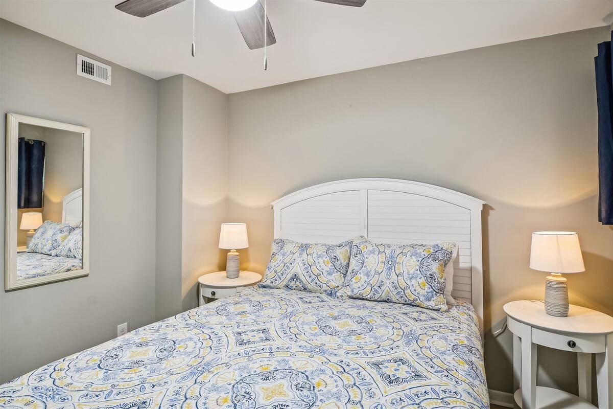 A bedroom with a bed, two bedside tables with lamps, a ceiling fan, a mirror, and a window with curtains.