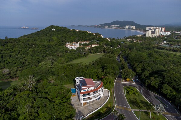 An aerial view of a coastal area with a prominent building featuring a curved roof and solar panels, surrounded by lush greenery and a winding road. - La Loma Suites (Ixtapa)