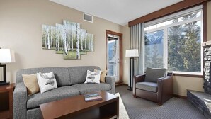 A modern living room with a grey sofa, a brown armchair, and a coffee table. There are three wall-mounted paintings of birch trees. A large window offers a view of a mountain and forest. - Beautiful Mtn Views-King Bed, Hot Tub (Canmore)