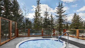 A hot tub with a wooden deck and glass walls, surrounded by trees and mountains. - Beautiful Mtn Views-King Bed, Hot Tub (Canmore)