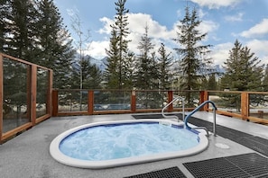 A hot tub with a wooden deck and glass walls, surrounded by trees and mountains.