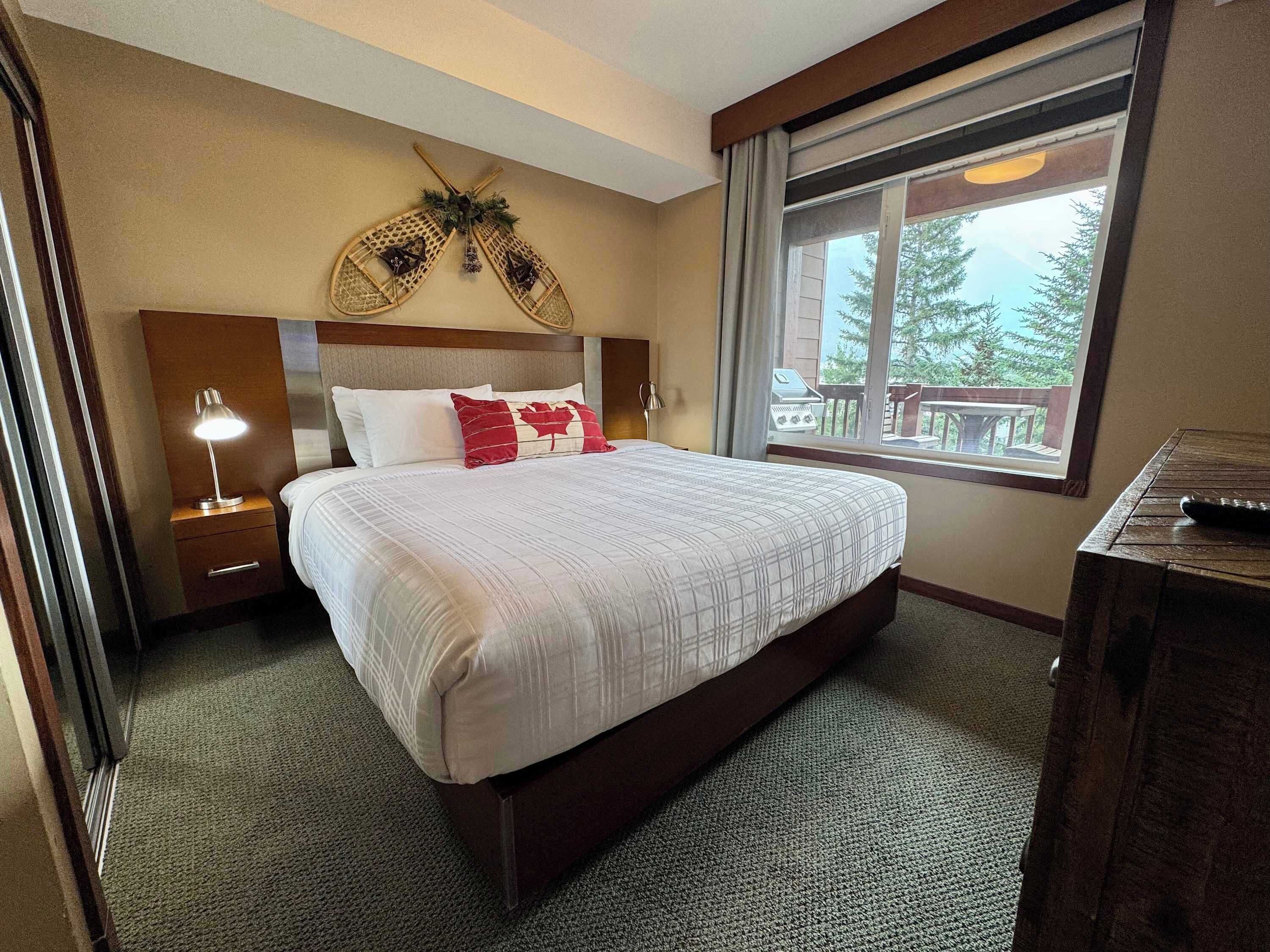 Een hotelkamer met een groot bed, een nachtkastje met lamp, een houten hoofdeinde, een raam met uitzicht op bomen en een houten dressoir.