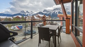Een balkon met rieten stoelen en een tafel, met uitzicht op een bergketen en een stad met sneeuw bedekte daken.