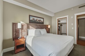 A hotel room with a large bed, a wooden headboard, a nightstand with a lamp, a wardrobe, and a painting on the wall. - Luxurious Corner Condo -Sunset Ridge (Canmore)