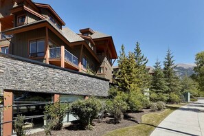 A multi-story building with a stone facade, large windows, and a well-maintained garden with various shrubs and trees. - Luxurious Corner Condo -Sunset Ridge (Canmore)