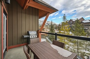 A balcony with a grill, wicker furniture, and a view of snow-covered buildings and trees. - Luxurious Corner Condo -Sunset Ridge (Canmore)