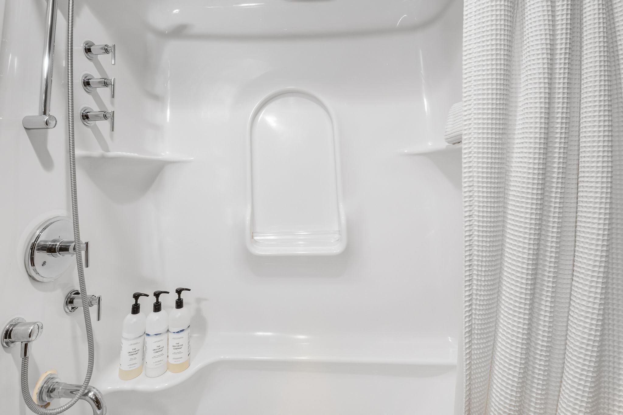 A white bathtub with a handheld showerhead, a built-in seat, and three bottles of shampoo.