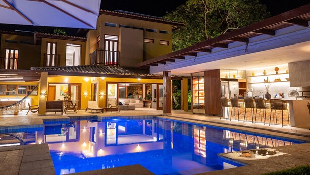 Un área de piscina exterior moderna con bar y asientos, rodeada por un edificio de dos pisos con ventanas grandes y un porche cubierto.