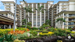 Un hotel a più piani con balconi, un giardino curato con piante tropicali e un cielo sereno.