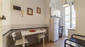 Una cucina compatta con un tavolo bianco, due sedie, un frigorifero e una finestra che si affaccia su edifici.