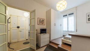 Una stanza con televisione su un mobile, una porta con maniglia, un quadro incorniciato appeso al muro e una finestra con persiane.