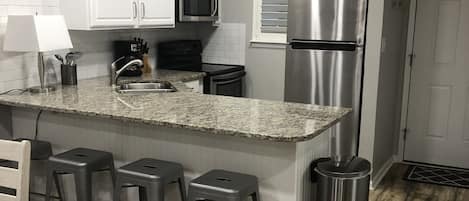 A kitchen with white cabinets, a stainless steel refrigerator, a microwave, a sink, and a bar stool area.