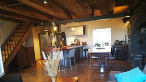 Een ruime woonkamer met houten vloer, een eettafel en een bank met blauwe kussens. Achtergrond toont een keuken met moderne apparatuur en een stenen open haard.