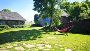 Een stenen pad dat leidt naar een hangstoel in een tuin met stenen muren en een blauwe deur.