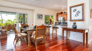 A spacious dining area with wicker chairs and a glass table, a kitchen with wooden cabinets, and a large window offering a view of greenery.