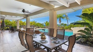 A covered outdoor dining area with a pool view, featuring a table set for four, chairs, and a ceiling fan.