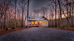 A cabin with a porch, a fire pit, and a gravel driveway surrounded by trees.