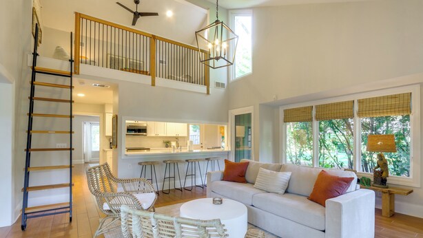 A spacious living room with a white sofa, wicker chairs, and a round coffee table. A wooden ladder leads to an upper level with a balcony. - Charming Lakefront Retreat in Palmetto Dunes! (Hilton Head Island)