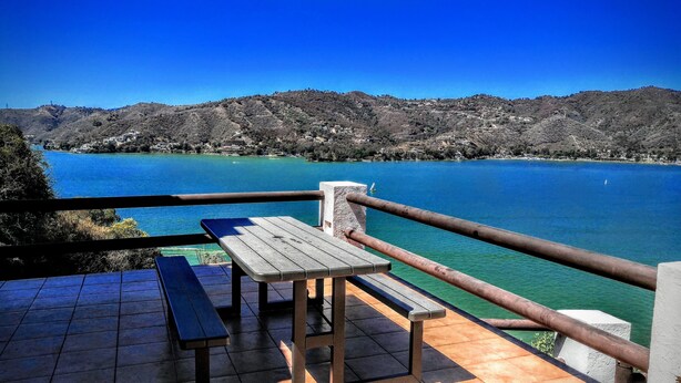Een terras met een tafel en banken met uitzicht op een meer met helderblauw water en een heuvelachtig landschap op de achtergrond.