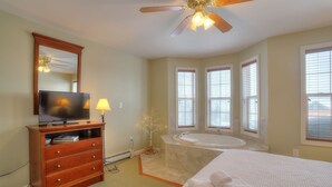 Une chambre d’hôtel comprenant un lit, une télévision, une commode en bois, une salle de bain avec baignoire et un ventilateur de plafond.