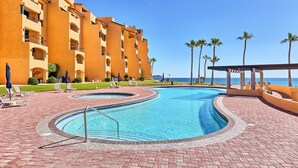 Une piscine à bord incurvé, entourée d’un chemin pavé en briques, et située à côté d’un immeuble à plusieurs étages doté de balcons et de palmiers.