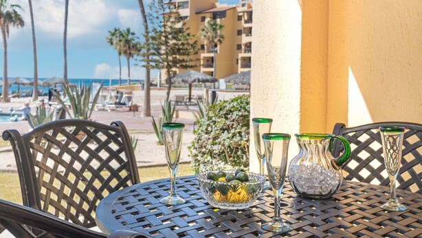 Une table sur laquelle se trouve un saladier en verre rempli de fruits, deux verres en cristal et une carafe, le tout posé sur une table en métal. En arrière-plan, on aperçoit une plage et des bâtiments d’hôtel.