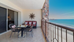A balcony with a table and chairs, a sofa with patterned cushions, and a decorative sun-like wall art. A stone wall and a metal railing are visible, with a view of the beach and ocean in the background. - Sonoran Sky 1403 Exquisite Spacious Ocean View Condo (Puerto Peñasco)