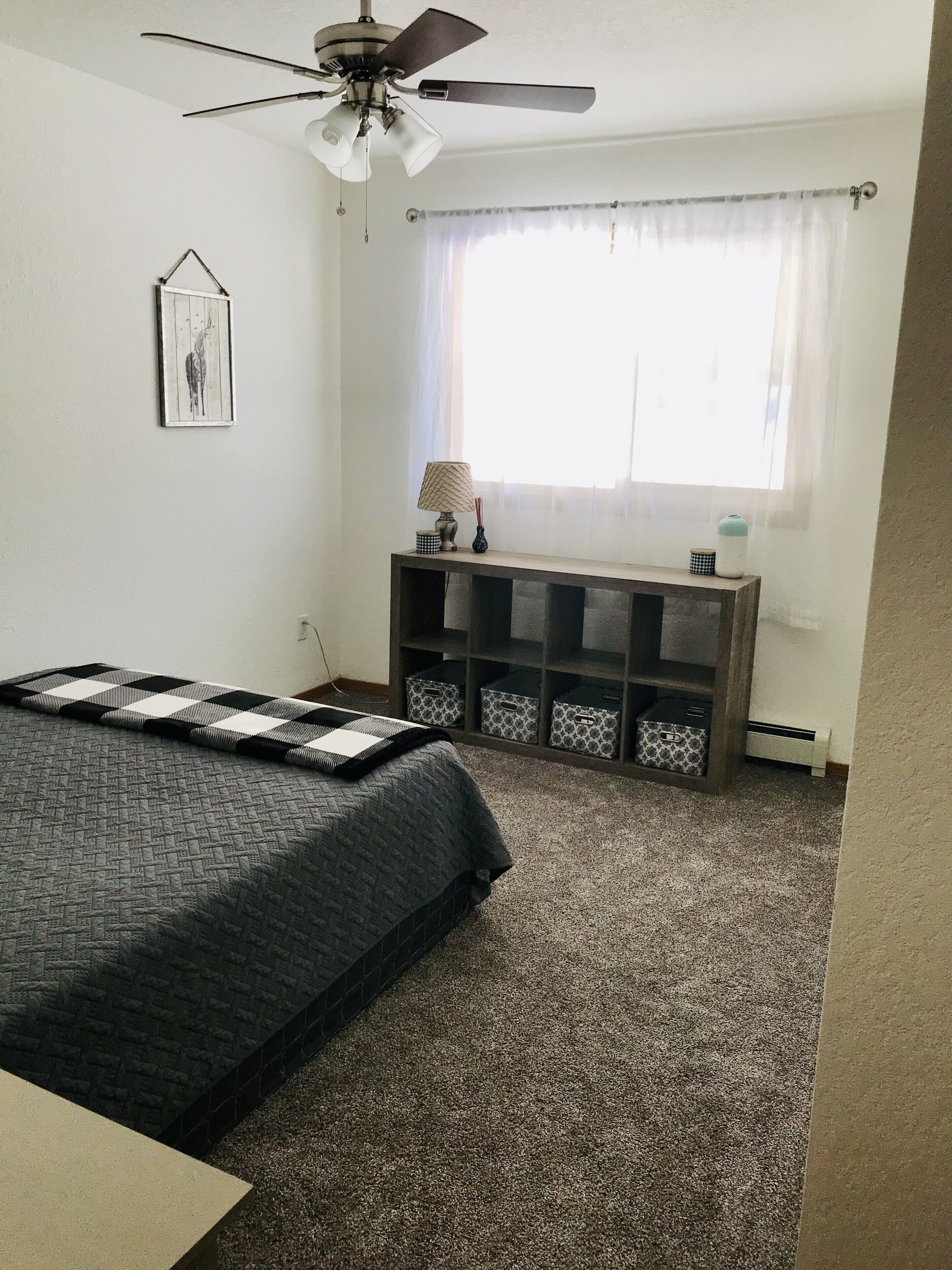 Un dormitorio con cama, ventilador de techo, ventana con cortinas, mesita de noche con lámpara y cajas de almacenamiento debajo de la ventana.