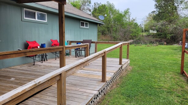 A wooden deck with a railing, a covered patio area with chairs and a table, and a grassy yard with trees in the background. - Tesla's lake Escape (Onalaska)