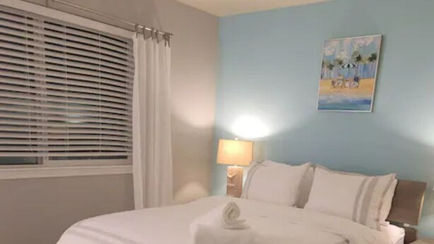Une chambre à coucher avec un lit, une table de chevet, une lampe, une fenêtre avec des stores et un tableau au mur.