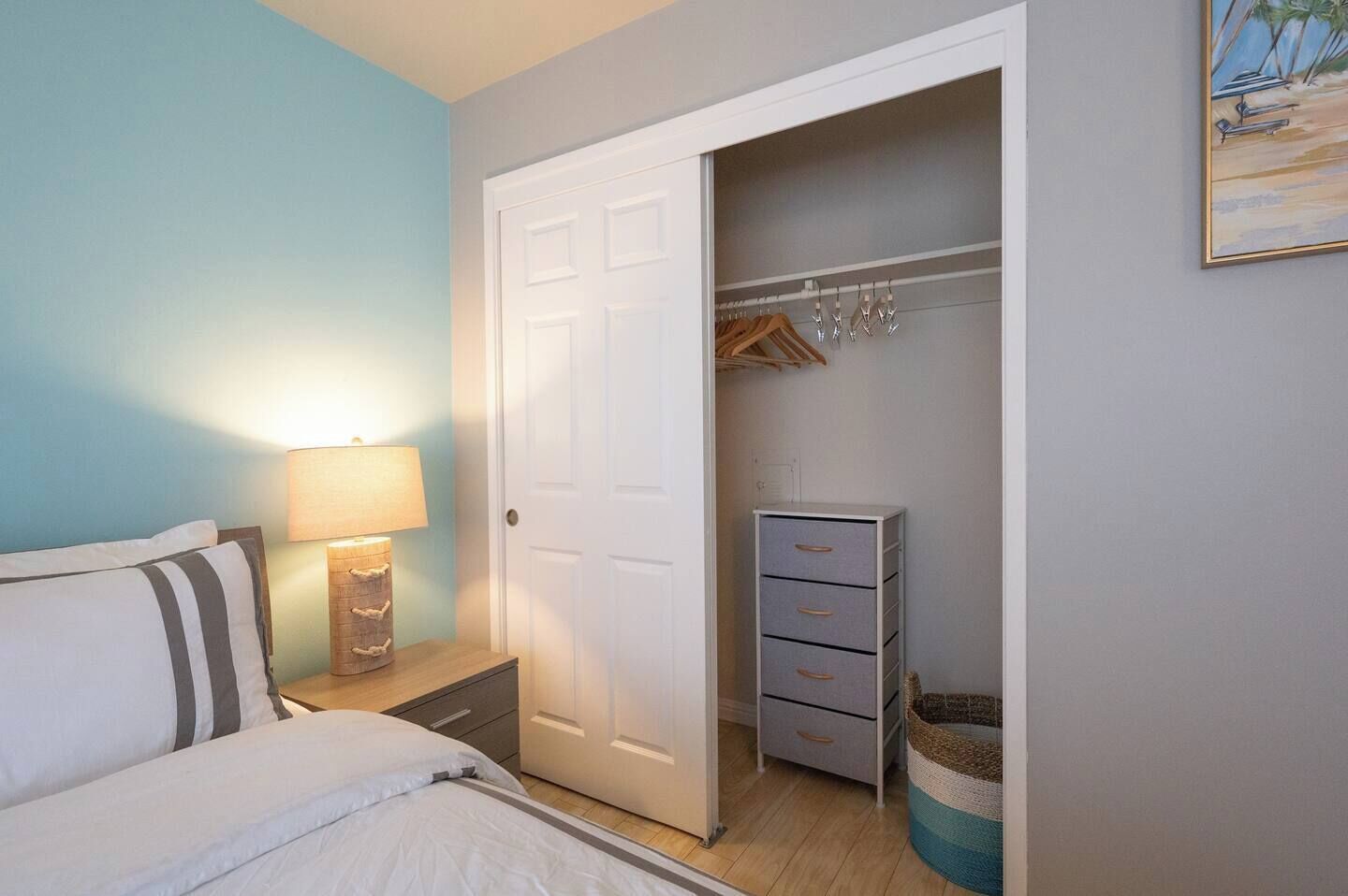 A bedroom with a bed, bedside lamp, and a closet with hanging clothes and storage drawers.