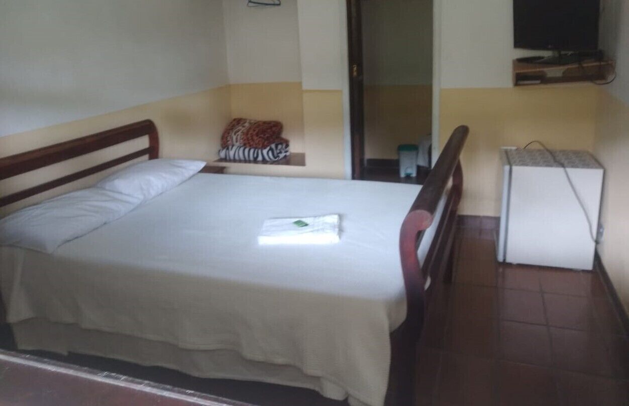 A single bed with a wooden headboard, a bedside table with a lamp, and a small shelf with a patterned pillow.