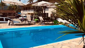 A swimming pool with lounge chairs and umbrellas, surrounded by palm trees and a building with a tiled roof. - Pousada Thamalu 228 (Paraty)