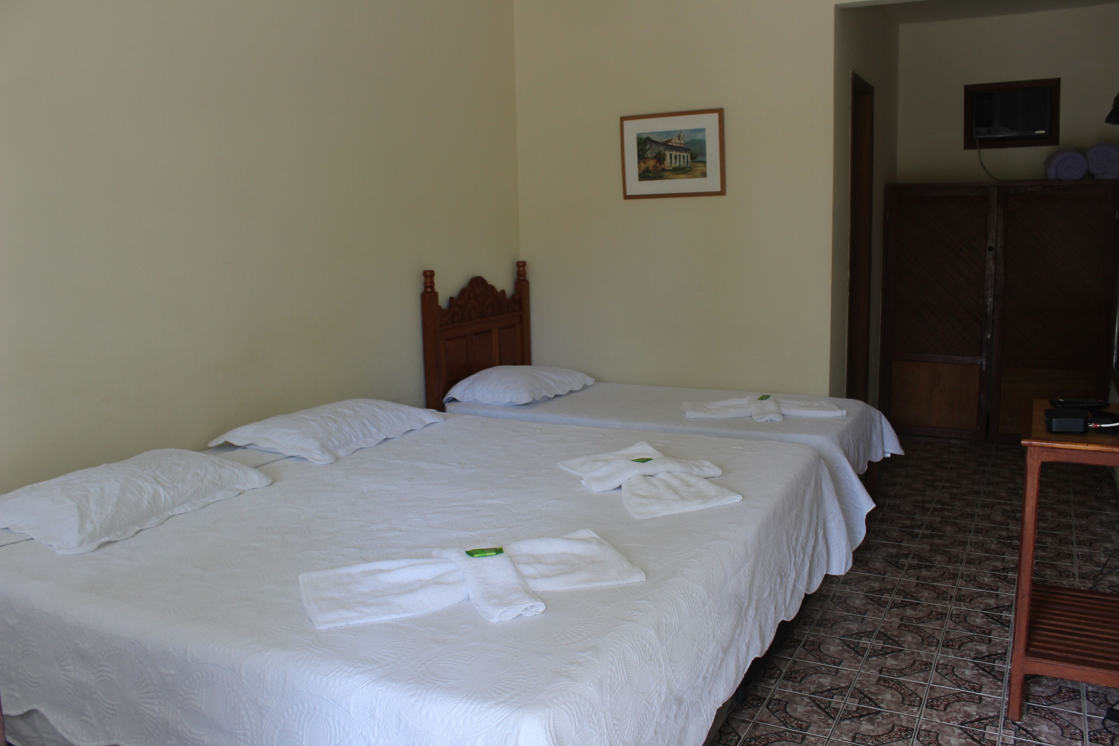 A hotel room with two beds, each with white linens and towels. There is a wooden headboard on one bed, a framed picture on the wall, and a television mounted above a wooden wardrobe.