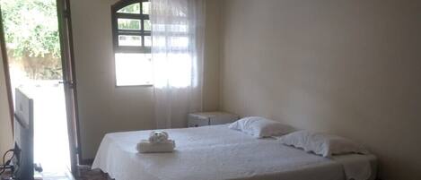 Una habitación sencilla y limpia con una cama, una mesita de noche pequeña y una ventana con cortinas.