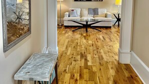 Un couloir au plancher de bois mène à un salon où se trouve un canapé blanc et un tableau accroché au mur.