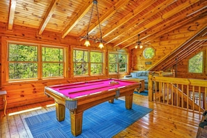 A wooden cabin with a pool table, large windows, and a staircase.