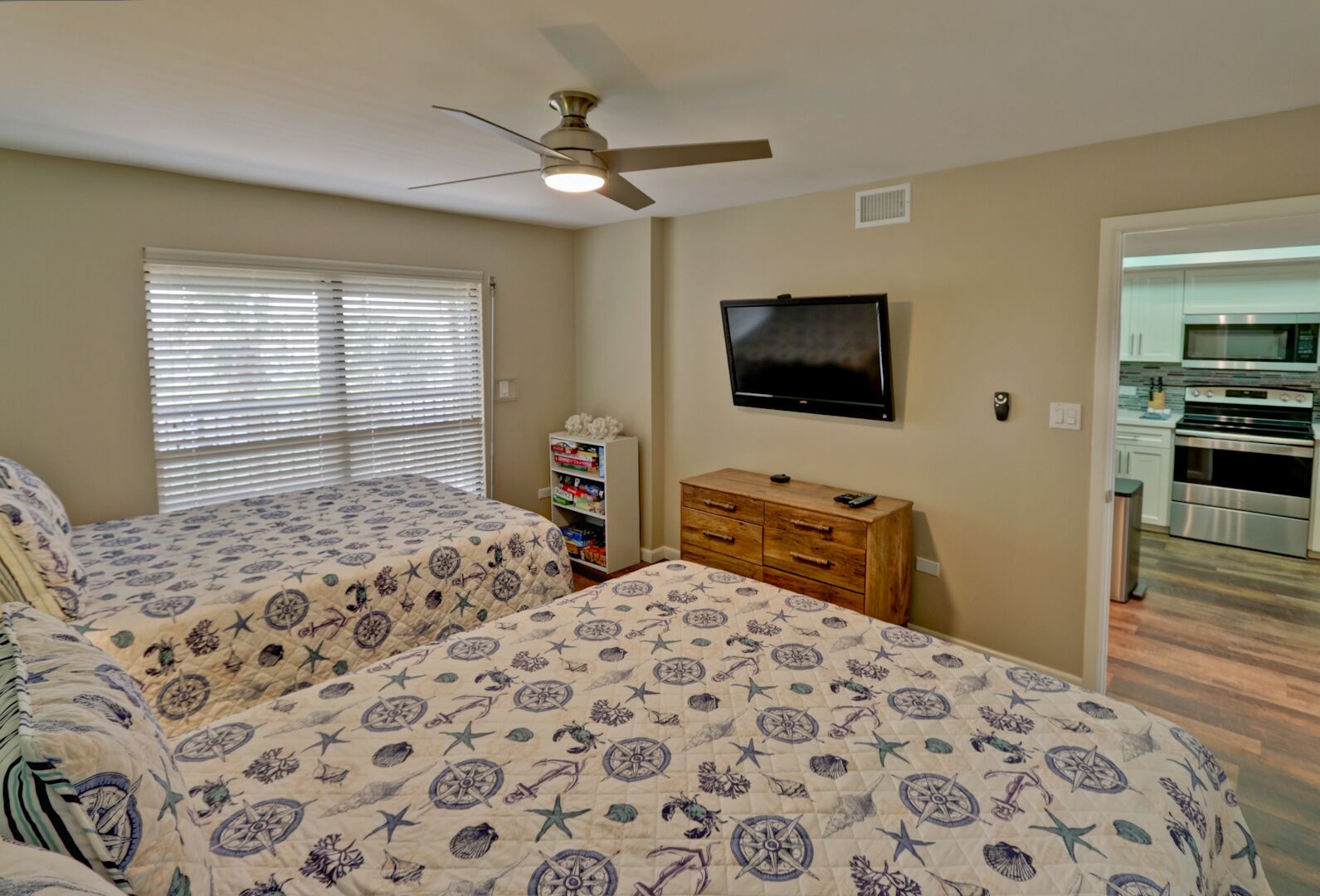 A bedroom with two beds, a ceiling fan, a TV, and a kitchenette in the background.