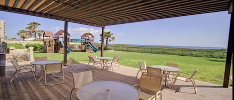 An outdoor patio with tables and chairs, a playground in the background, and a view of the ocean.