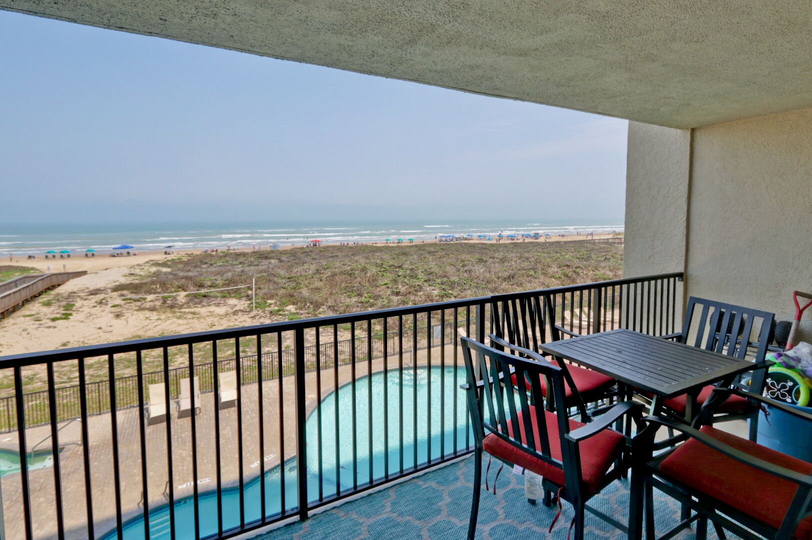 A balcony with a pool, chairs, and a table overlooking a beach and ocean.