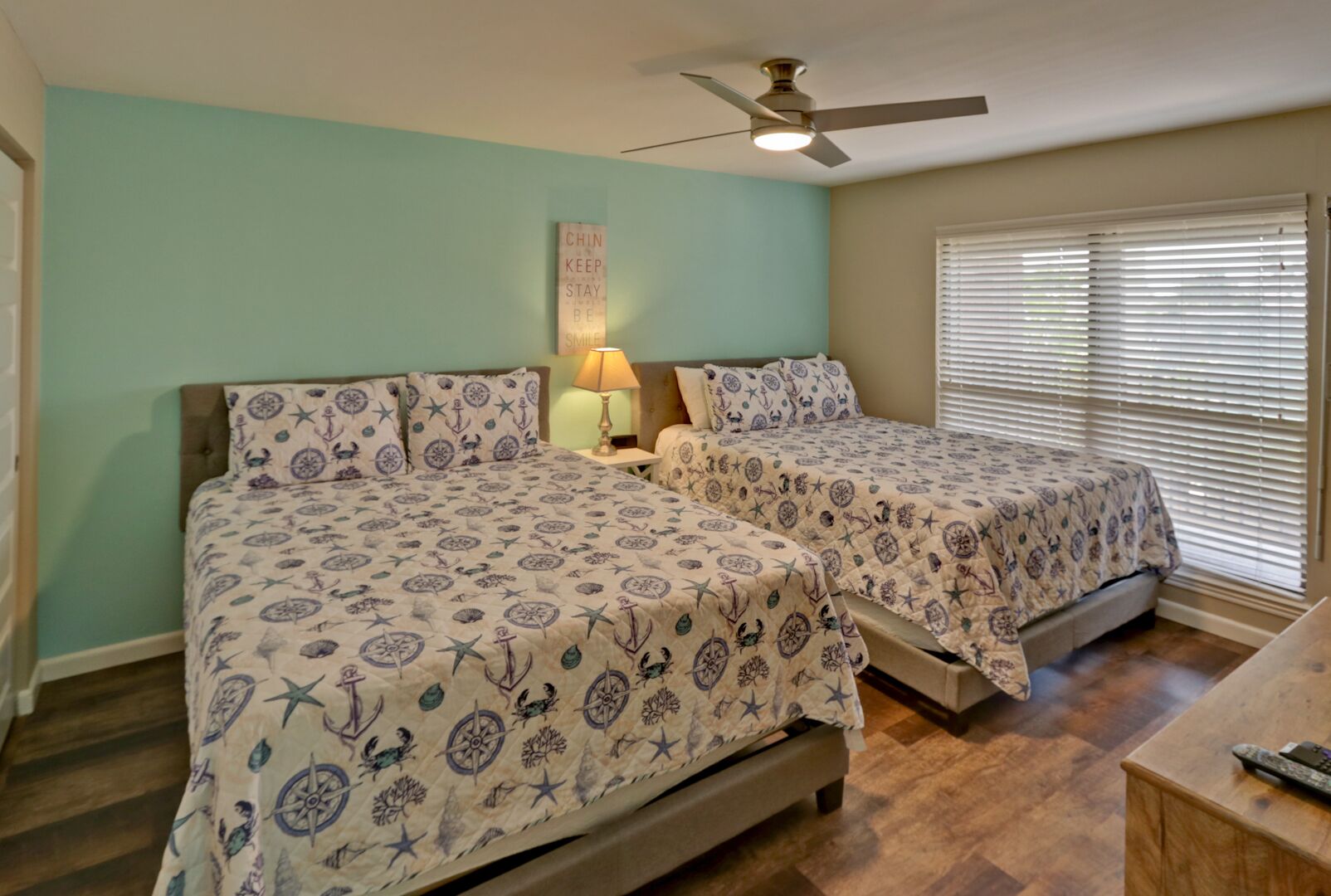 A bedroom with two beds, a ceiling fan, a nightstand with a lamp, and a window with blinds.