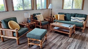 Quarto com piso de madeira, cadeiras e sofá estofados em verde, uma mesa de centro de vidro e uma luminária. Janelas com molduras de madeira oferecem vista para o verde lá fora.