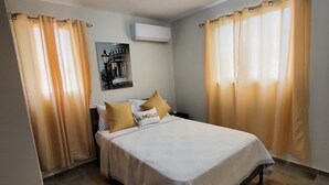 Un dormitorio con cama, cortinas, unidad de aire acondicionado y un cuadro colgado en la pared.