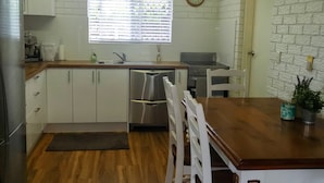 A compact kitchen with white cabinets, a stainless steel refrigerator, a sink, a stove, and a dining table with white chairs. - Jack's Shack - Burrum Heads - Relax, fish and swim (Burrum Heads)